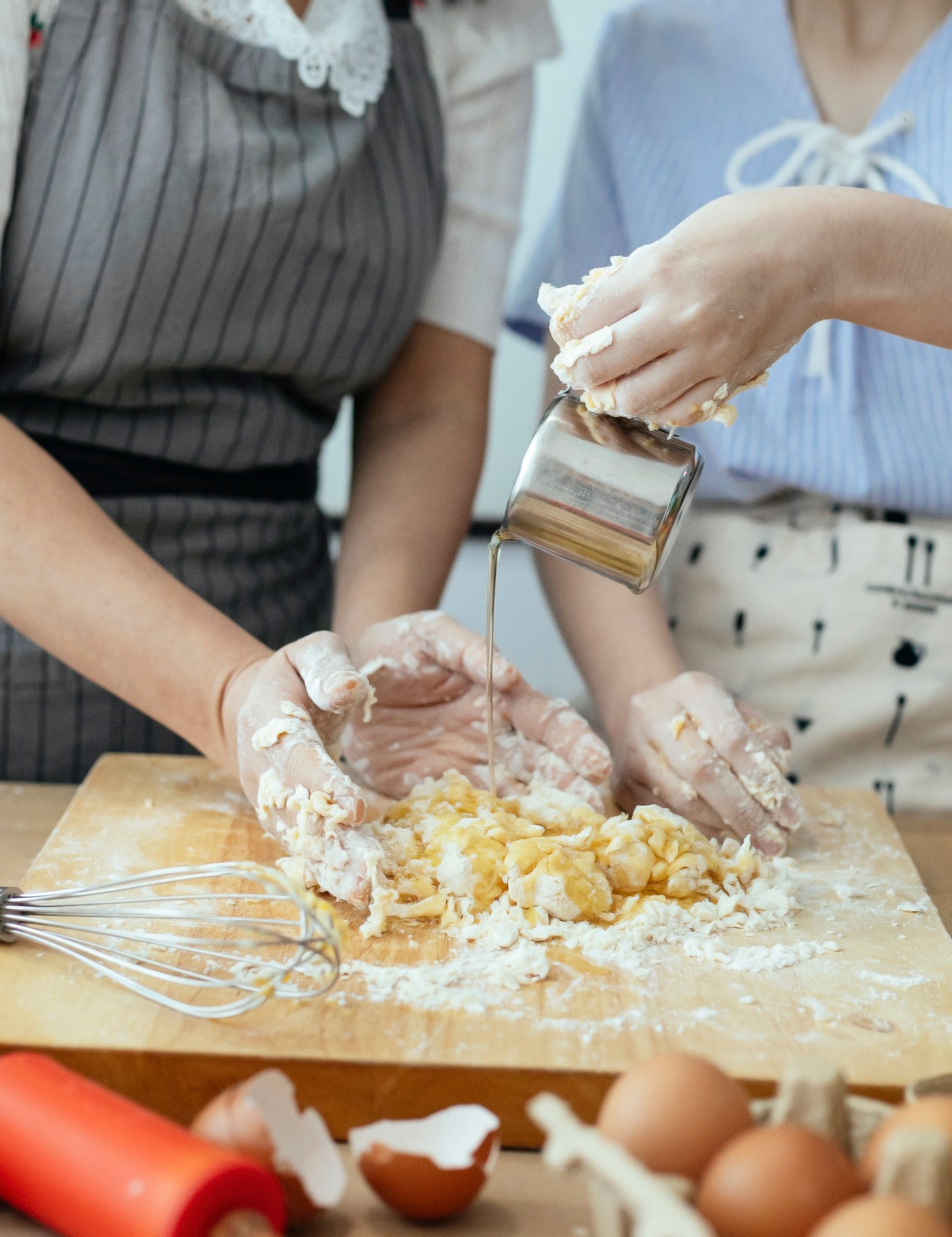 Crop anonymous housewife in apron preparing fresh dough from flour and eggs while daughter pouring cold water from jug into dough